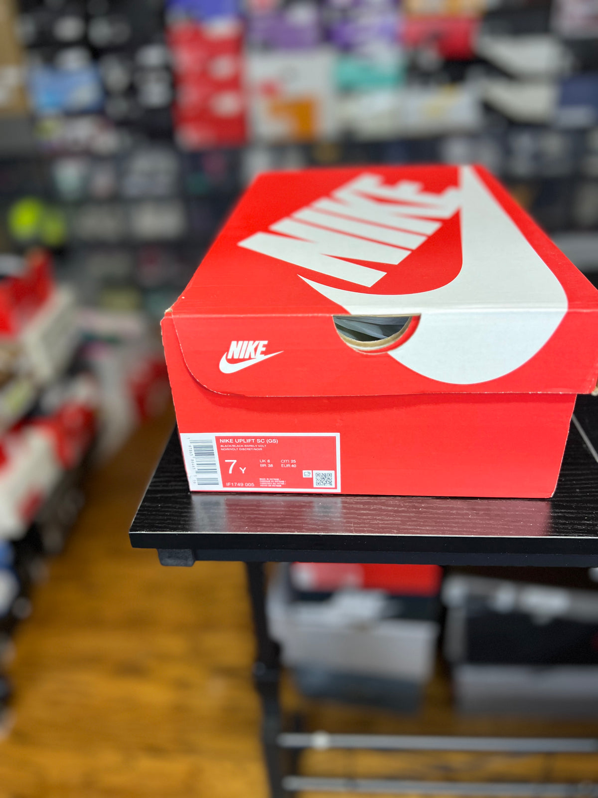 A red Nike shoebox with a large white swoosh and logo sits on a black table in a store setting with blurred shelves in the background.