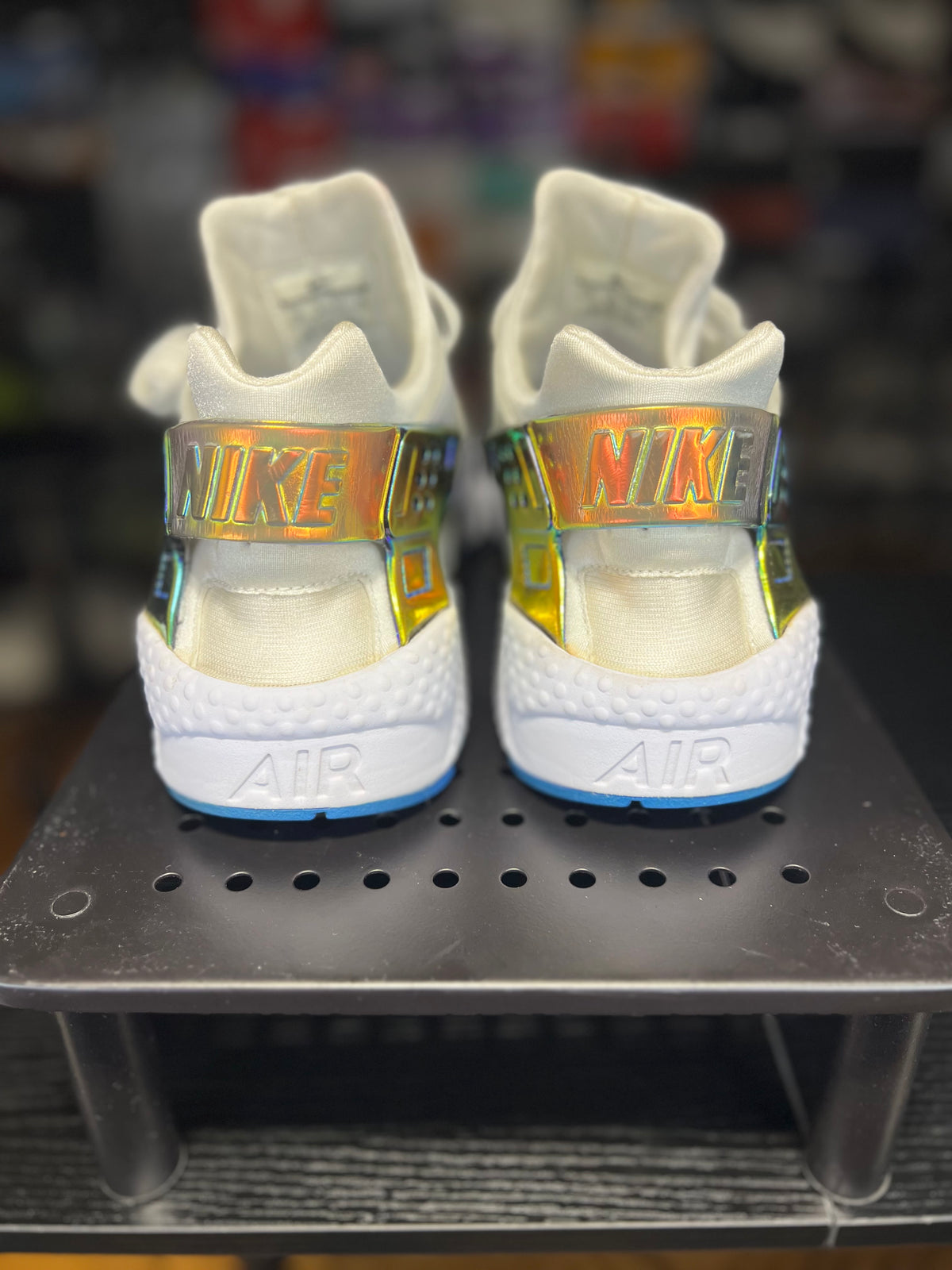 A pair of Nike high-top sneakers shown from the back, featuring white uppers with metallic holographic Nike straps and white midsoles with blue outsoles, displayed on a black metal stand.