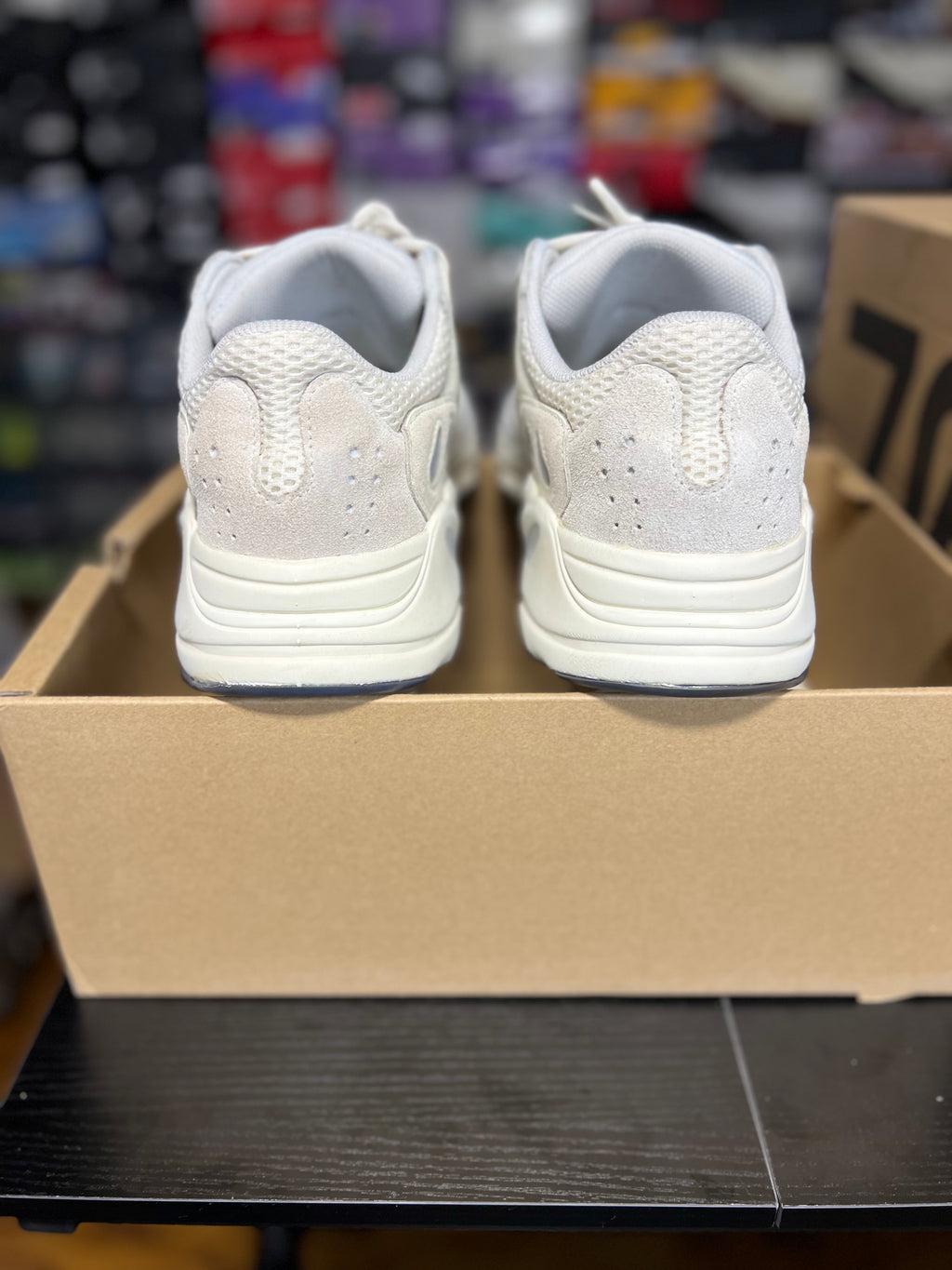 A pair of white athletic sneakers shown from the back, resting on top of a brown cardboard shoebox in a store setting.
