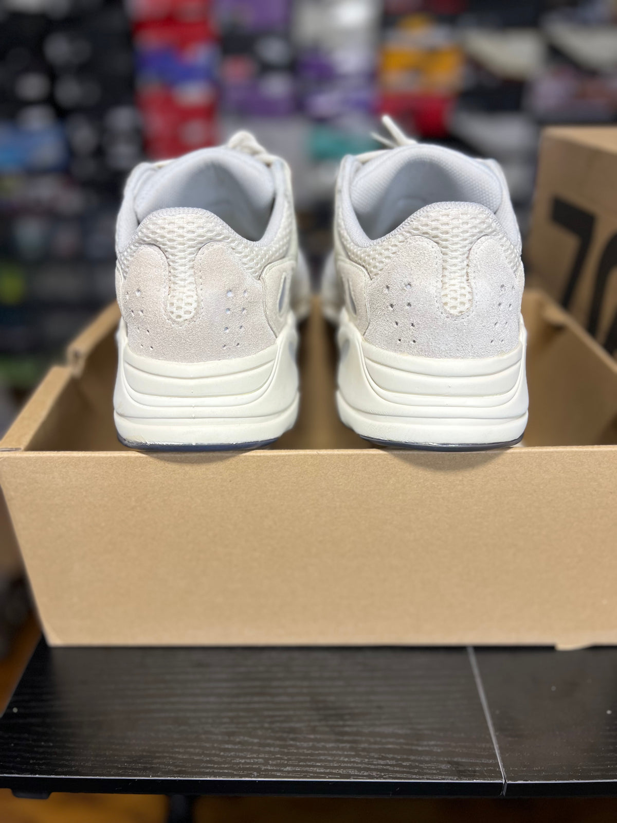 A pair of white athletic sneakers shown from the back, resting on top of a brown cardboard shoebox in a store setting.