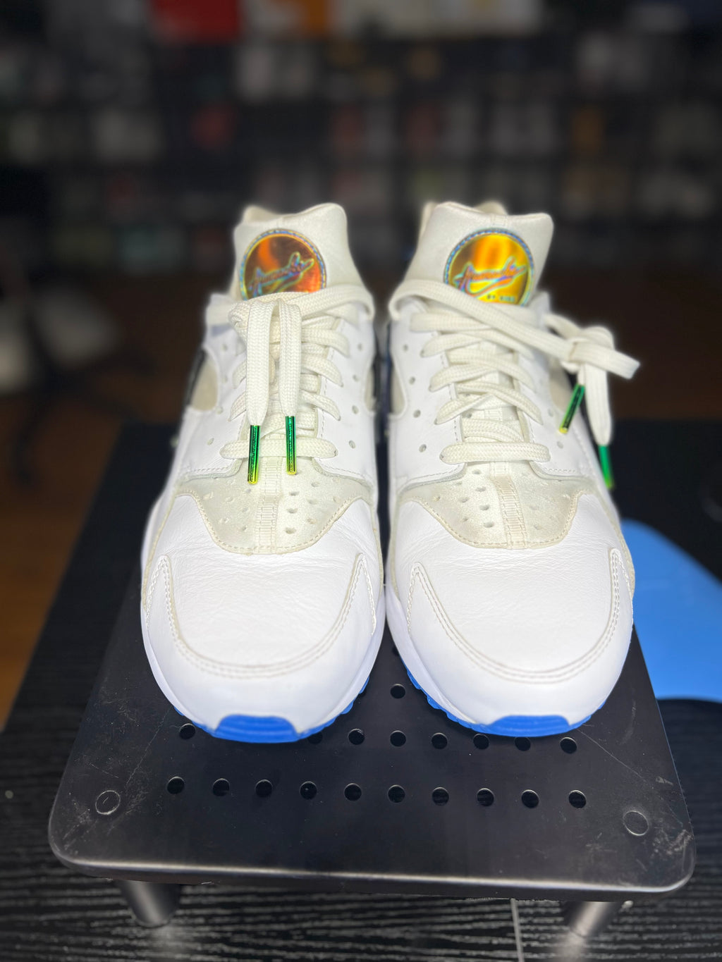 A pair of white Nike Air Max 90 sneakers with green and blue accents, displayed on a black metal stand.
