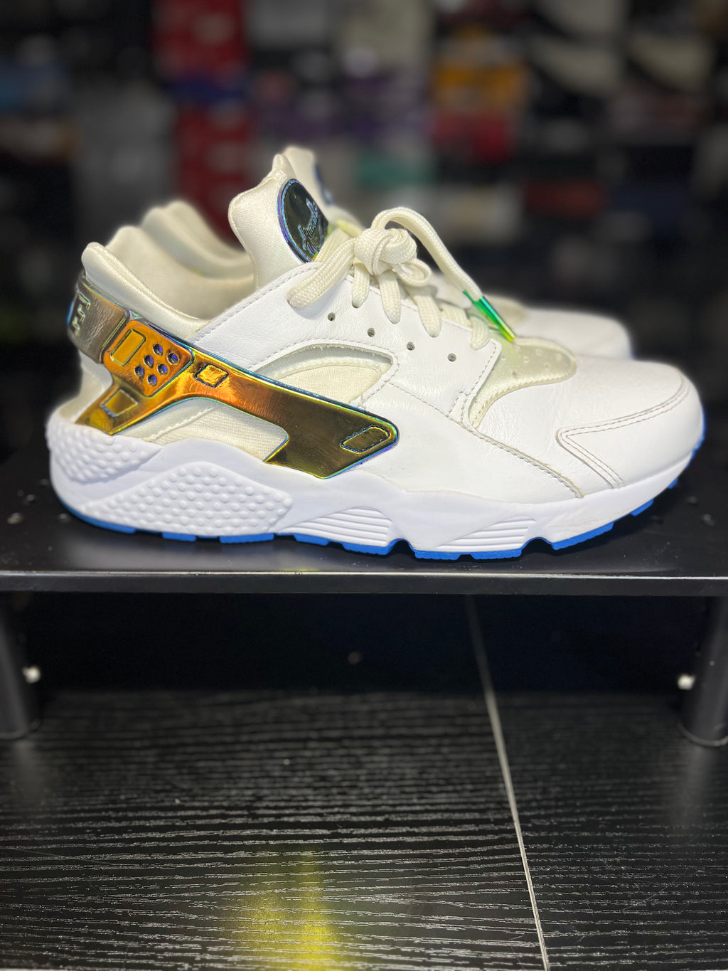 A white Nike Air Max sneaker with metallic gold and iridescent accents, displayed on a black shelf.
