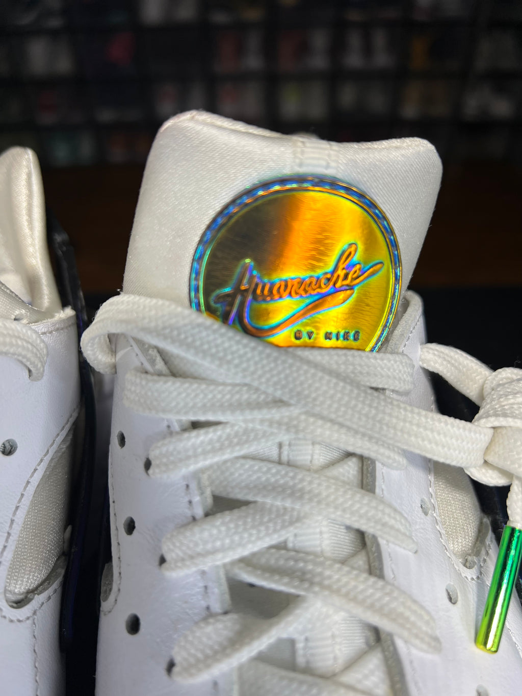 Close-up of a white sneaker with white laces and a prominent holographic circular logo patch on the tongue that reads 'Punisher' and 'BY NIKE'.
