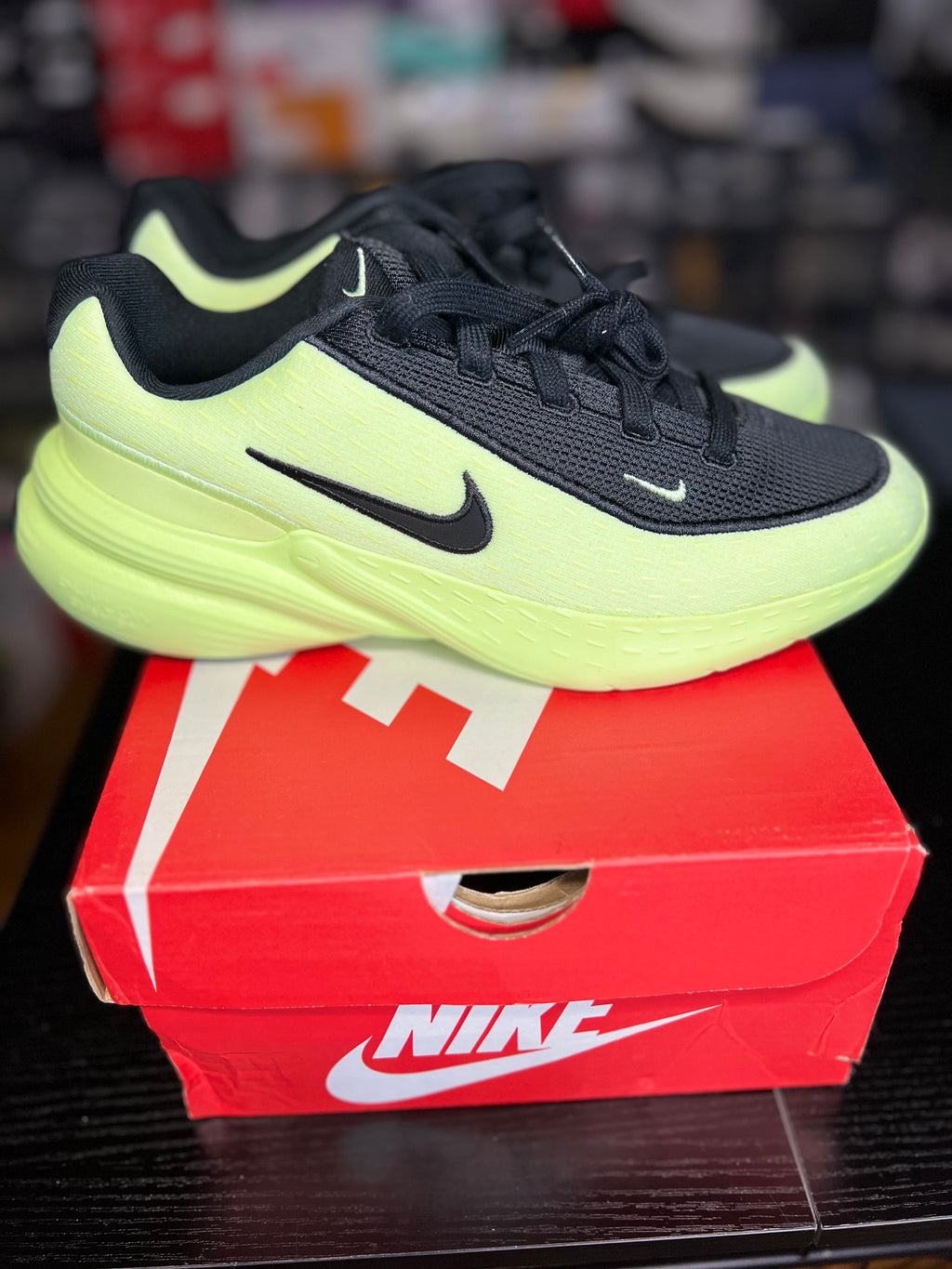A pair of Nike running shoes in black and neon yellow-green, displayed on top of a red Nike shoebox on a dark wooden surface.