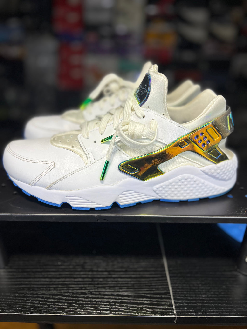A pair of white Nike Air Max sneakers with a metallic gold and green side stripe, displayed on a black shelf.
