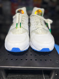 A pair of white low-top sneakers with bright blue soles and green-and-white laces, displayed on a black perforated shelf.