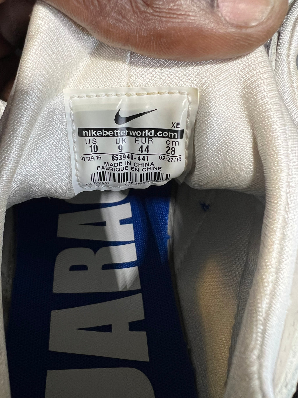 Close-up of the inside collar of a Nike shoe showing the size and model tag, with a finger holding the fabric open. The tag reads nikebetterworld.