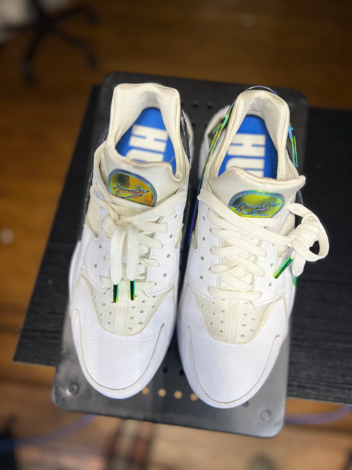 A pair of white high-top sneakers with green and blue accents, displayed on a black metal stand over a wooden floor.