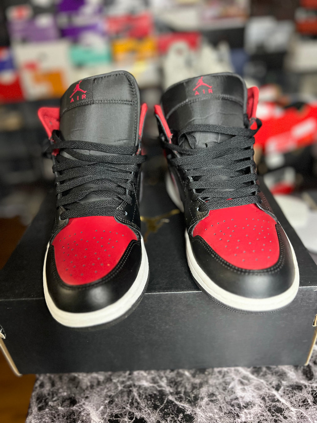 A pair of black and red high-top Jordan 1’s displayed on top of a black shoebox, with a blurred background of shelves containing various shoeboxes. The sneakers feature a black leather upper with vibrant red suede overlays on the toe boxes and tongue, black laces, and a white midsole.